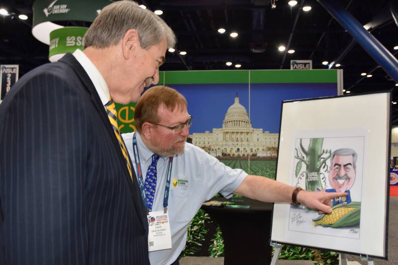 Dave Blower, news and media relations manager of the Indiana Soybean Alliance, the Indiana Corn Marketing Council and the Indiana Corn Growers Association, points out the artwork created by Gary Varvel, former editorial cartoonist for The Indianapolis Star, depicting Ted McKinney (left) in a Purdue Boilermakers tie and iconic FFA blue jacket and recognizing his efforts to promote global exports as the U.S. Department of Agriculture under secretary for trade and foreign agricultural affairs.